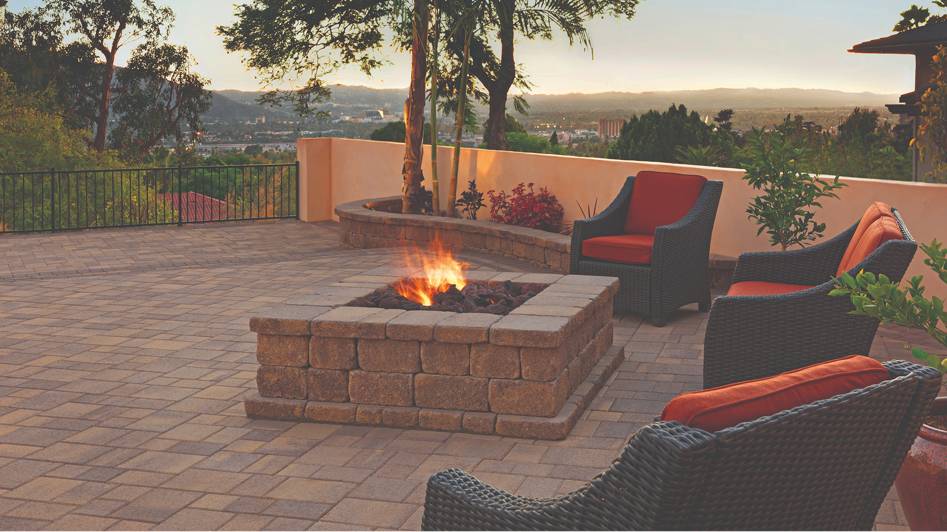 Elevated patio at sunset featuring a Sand-Stone-Mocha Courtyard Stone surface installed in an ashlar pattern with a solid Mocha Holland border. A square fire pit and curved planter wall are built with Stonewall® II in Sand-Stone-Mocha. Wicker lounge chairs with orange cushions surround the fire pit, while lush plants and a scenic valley view complete the relaxing outdoor atmosphere.