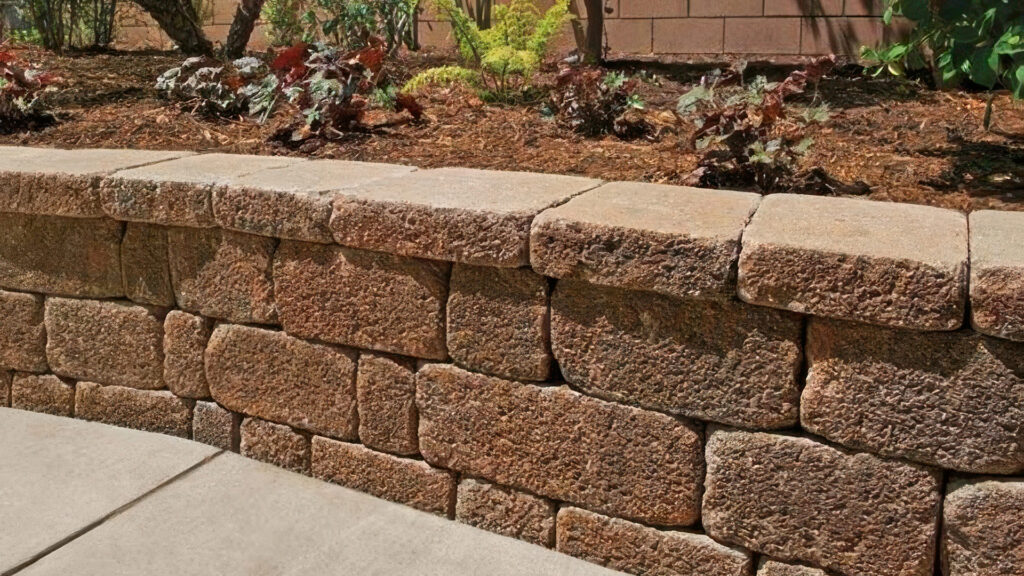 Planter wall constructed with Cream-Brown-Charcoal Stonewall® II blocks featuring a tumbled, textured finish. The wall separates a concrete patio from a mulched garden bed planted with decorative foliage and small shrubs in a residential backyard setting.