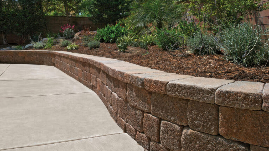 Gently curved planter wall constructed with Cream-Brown-Charcoal Stonewall® II blocks, separating a lush garden bed from a smooth concrete patio. The garden bed is filled with mulch and various drought-tolerant plants and shrubs, framed by a shaded backyard environment.