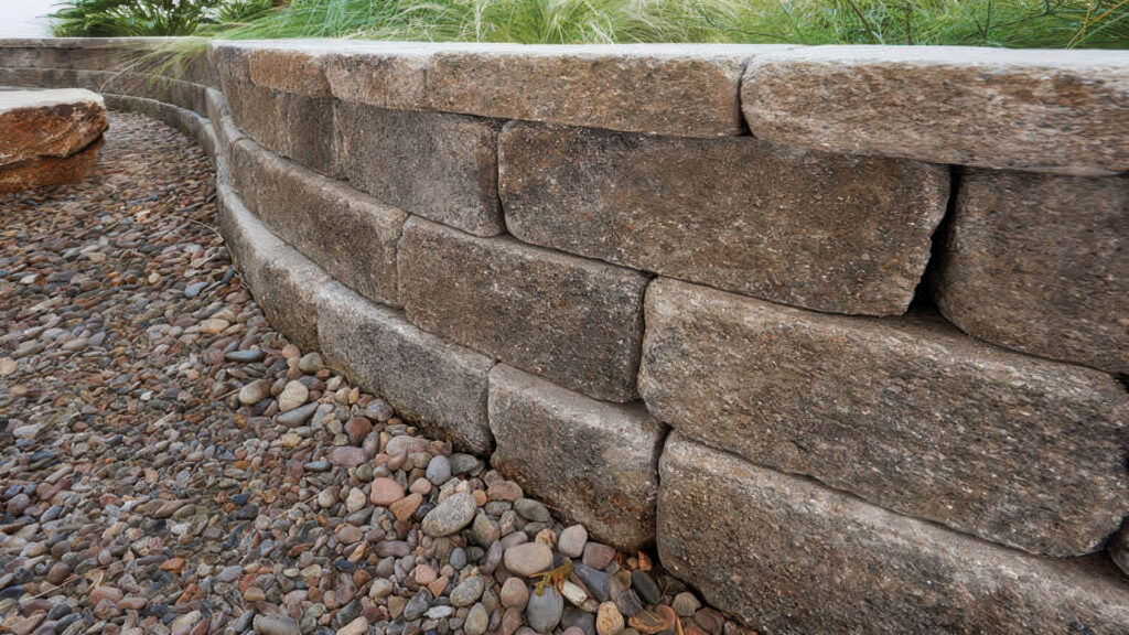 Close-up view of a curved planter wall built with Gray-Moss-Charcoal Stonewall® II blocks. The wall features a textured, tumbled appearance and borders a dry riverbed filled with smooth, multicolored pebbles. Ornamental grasses and foliage are planted above the wall.