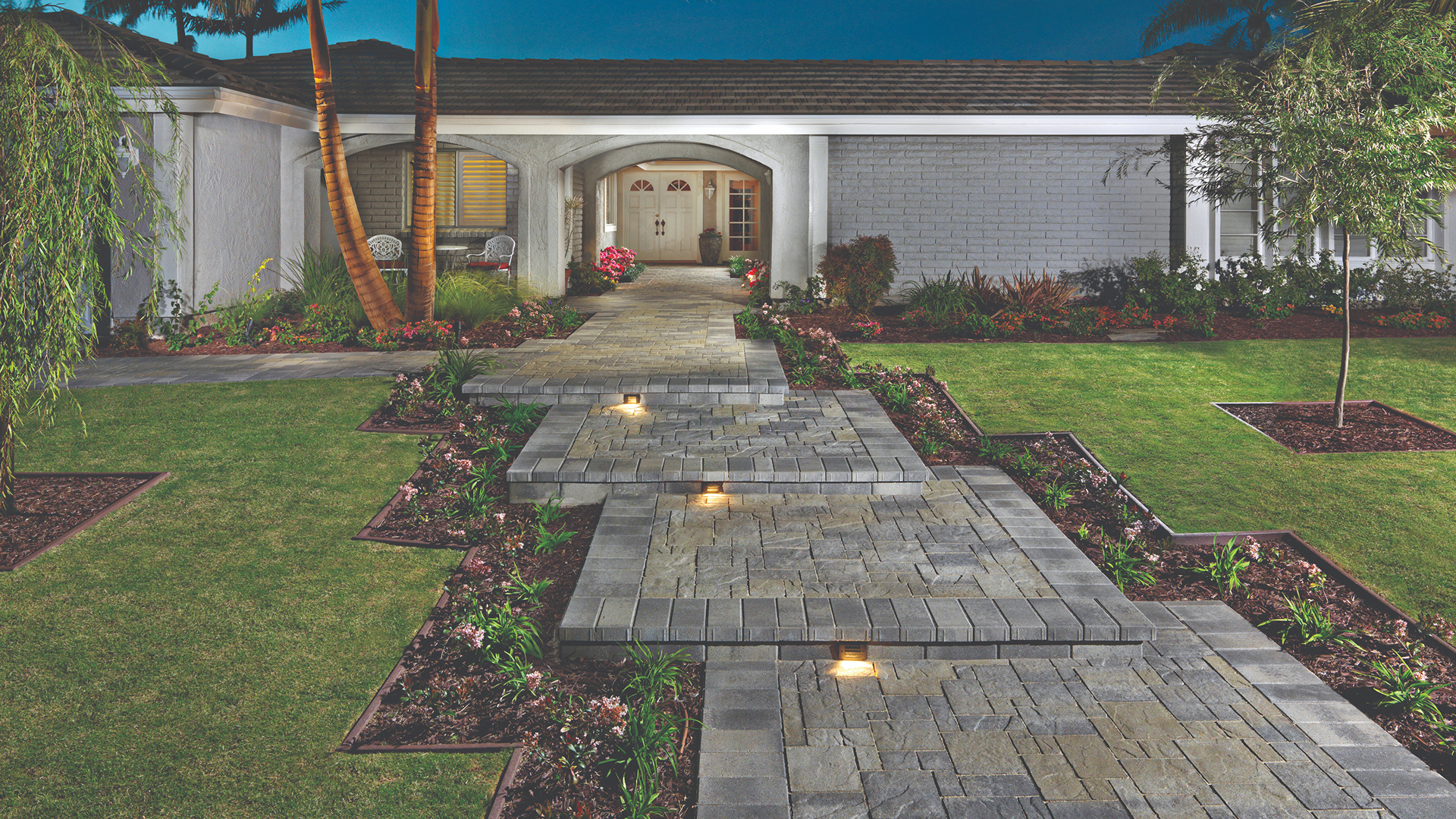 Heartland paving stones in a Gray-Moss-Charcoal blend arranged in an ashlar pattern, forming a modern walkway leading to a front entrance. The path features a 6x12 Gray-Charcoal border and integrated step lights, framed by manicured lawns and lush garden beds.