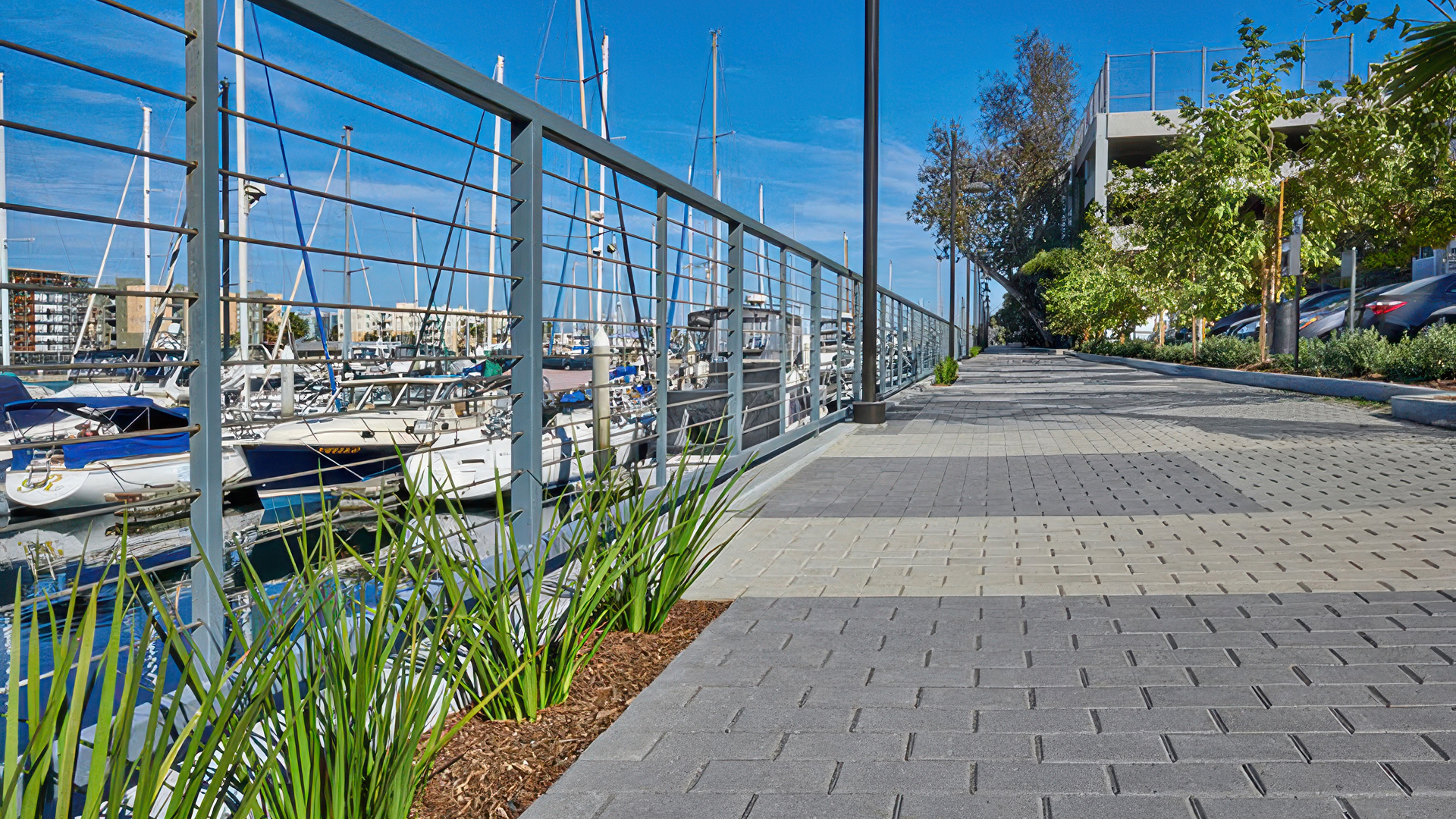 Waterfront pedestrian walkway next to a marina with sailboats docked behind a metal railing. The path is paved with Holland pavers in Light Charcoal, Midnight, and Gray (special order colors), arranged in a running bond pattern. The pavers create a striped visual effect that guides foot traffic, bordered by mulch beds with ornamental grasses and a tree-lined parking area on the right.
