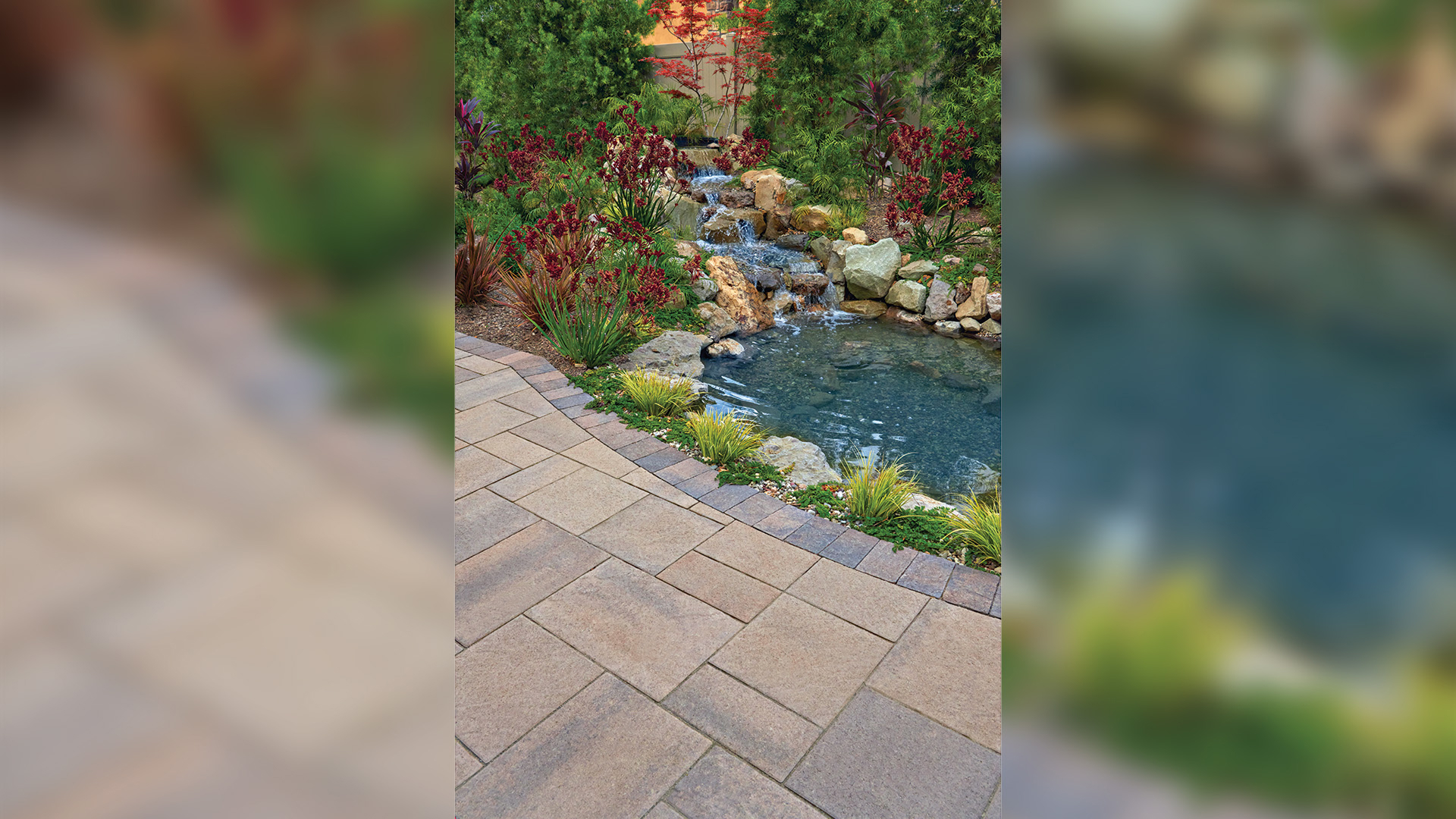 Outdoor patio paved with Pavilion I and II pavers in Dark-Gray-Copper-Charcoal, arranged in a random ashlar pattern. The border features Sand-Stone-Mocha Slate Stone 6x9 pavers in a soldier course. Adjacent to the patio is a landscaped koi pond with a cascading waterfall and vibrant plantings.