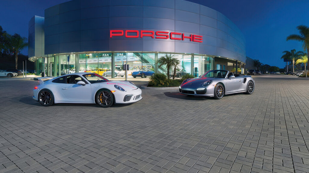 Two Porsche sports cars are parked in front of a modern Porsche dealership at dusk. The foreground features a clean installation of Permeable Holland pavers in Gray and Charcoal, arranged in a basketweave pattern. The sleek pavers complement the luxury setting, with palm trees and glowing dealership lights reflecting off the curved glass building.