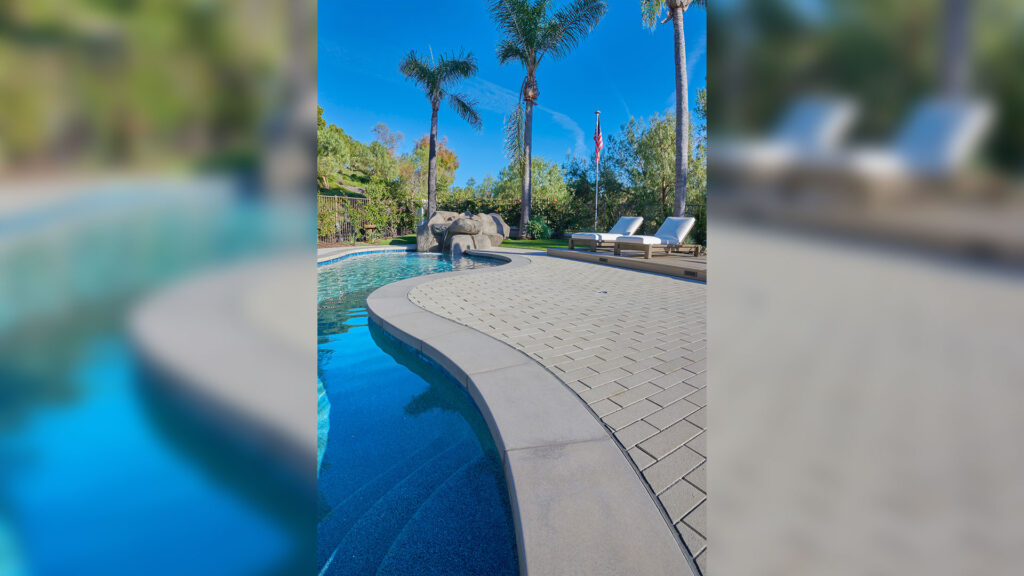 80mm 4x12 pavers in Gray, installed in a running bond pattern, create a curved pool deck surrounding a freeform swimming pool. The contemporary outdoor space features clean paving lines, palm trees, lounge chairs, natural rock accents, and lush landscaping under a clear blue sky.