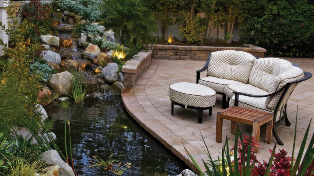 Sand-Stone-Mocha Rustic Wall Stone surrounds a tranquil pond with lush landscaping and a cascading waterfall. The patio features Sand-Stone Castle Cobble II (tumbled) pavers in an ashlar layout, bordered by a solid band of Sand-Stone Estate Cobble II (tumbled), framing a cozy seating area with curved retaining walls.