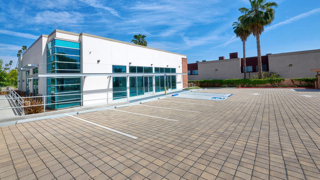 Accessible parking lot in front of a modern commercial building with large blue-tinted windows and palm trees in the background. The pavement is Sand-Stone-Mocha SF Rima pavers installed in a clean stack bond pattern, designed for both visual appeal and stormwater permeability.