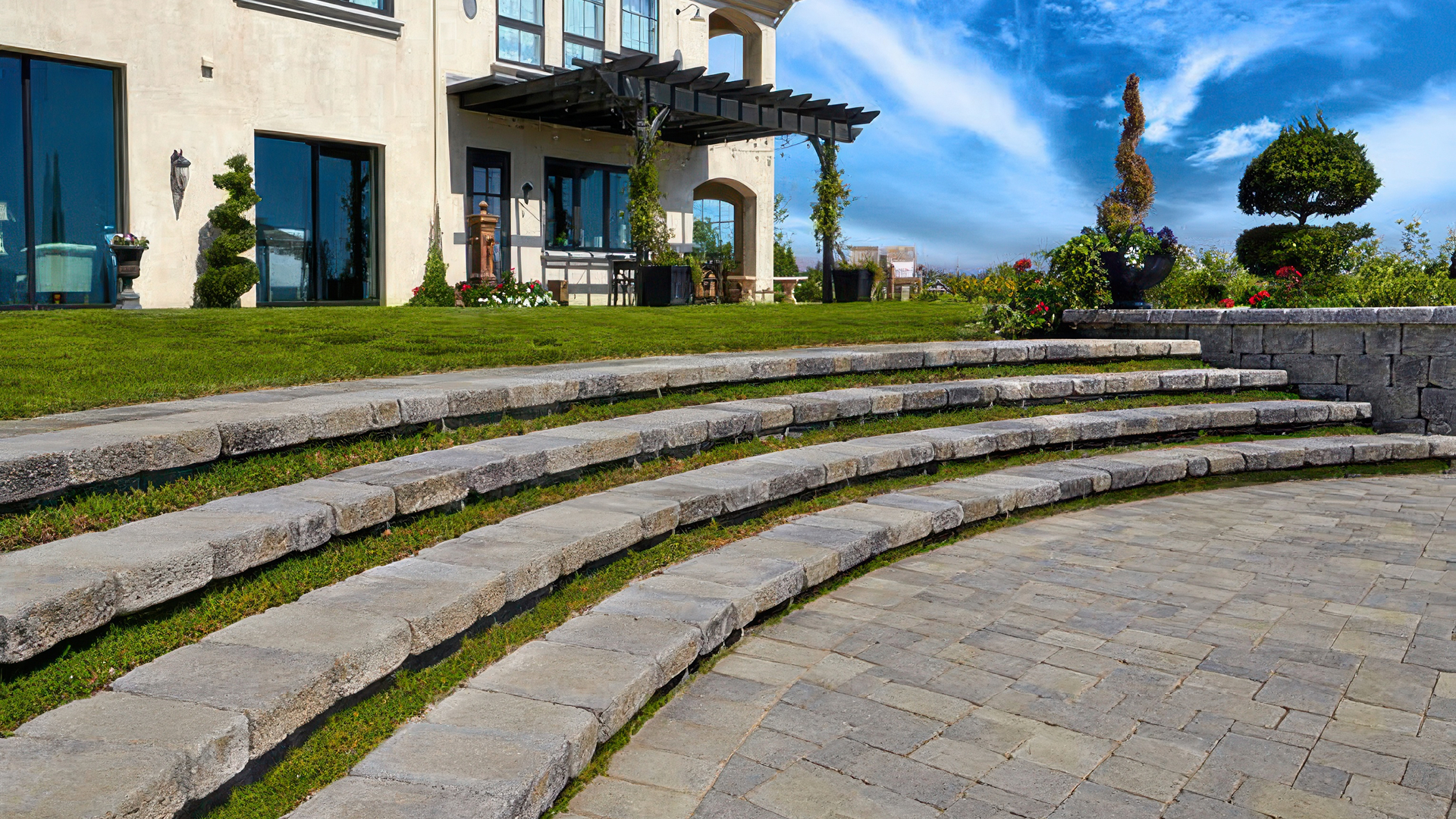 Outdoor tiered landscape with curved steps featuring Stonewall Caps in Gray-Moss-Charcoal used as treads and Stonewall® II Blocks for the risers and planter wall. The lower patio area is paved with Castle Cobble I and II (distressed) in Gray-Moss-Charcoal, installed in a random ashlar pattern. The scene is framed by manicured grass, colorful flowers, and topiary trees in a formal garden setting.