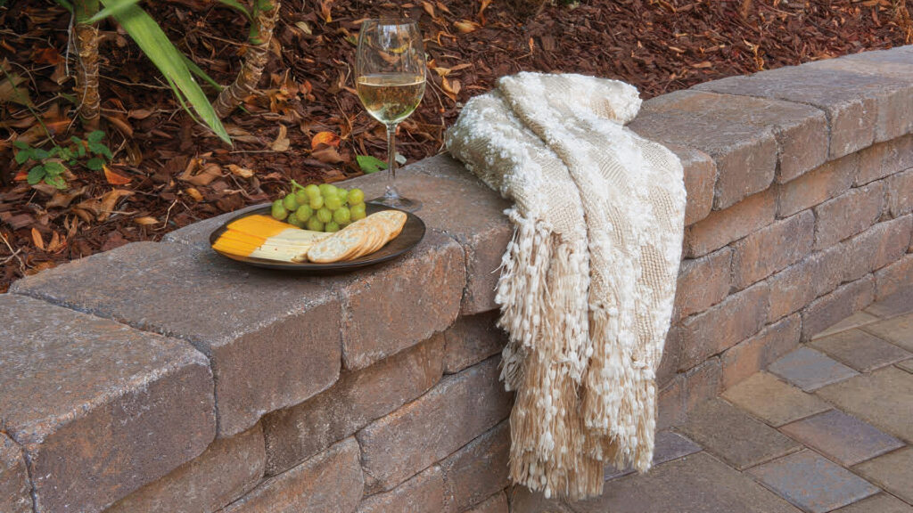 Close-up of a Tuscan Rustic Wall used as seating or a planter edge. A soft textured throw blanket is draped over the wall, with a plate of cheese, crackers, and grapes beside a glass of white wine, creating a cozy and inviting outdoor setting. The surrounding ground is covered with bark mulch and pavers.