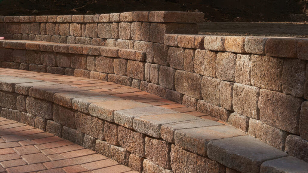 Tuscan Stonewall® II retaining wall with integrated tiered seating. The wall’s textured finish and natural blend of warm tones complement the curved design, offering both functional support and a built-in seating solution along the paved patio.
