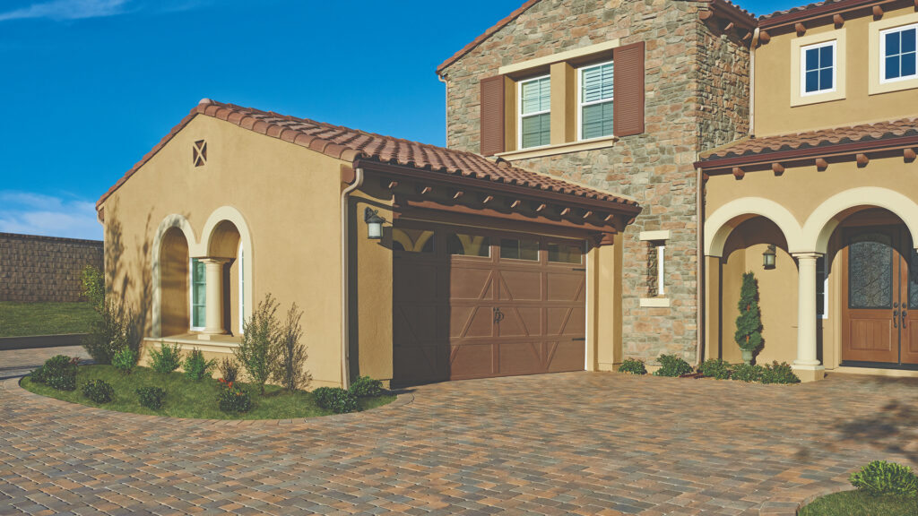 Close-up view of a driveway paved with Antique Cobble I and II in Tuscan blend, installed in a combination herringbone pattern. The Mediterranean-style home features warm stucco walls, arched entryways, brown garage doors, and stone façade accents with neatly manicured landscaping.