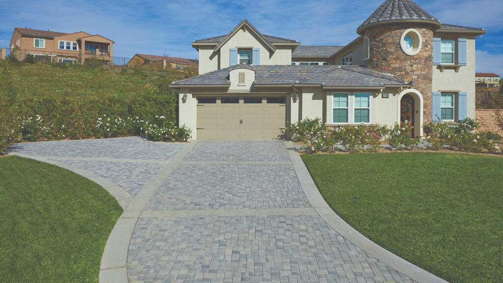 Residential driveway paved with Antique Cobble I & II in Gray–Charcoal blend, installed in a combination herringbone pattern. The home features light stucco siding, a turret-style stone accent, blue shutters, and manicured landscaping with rose bushes and lush green lawns.