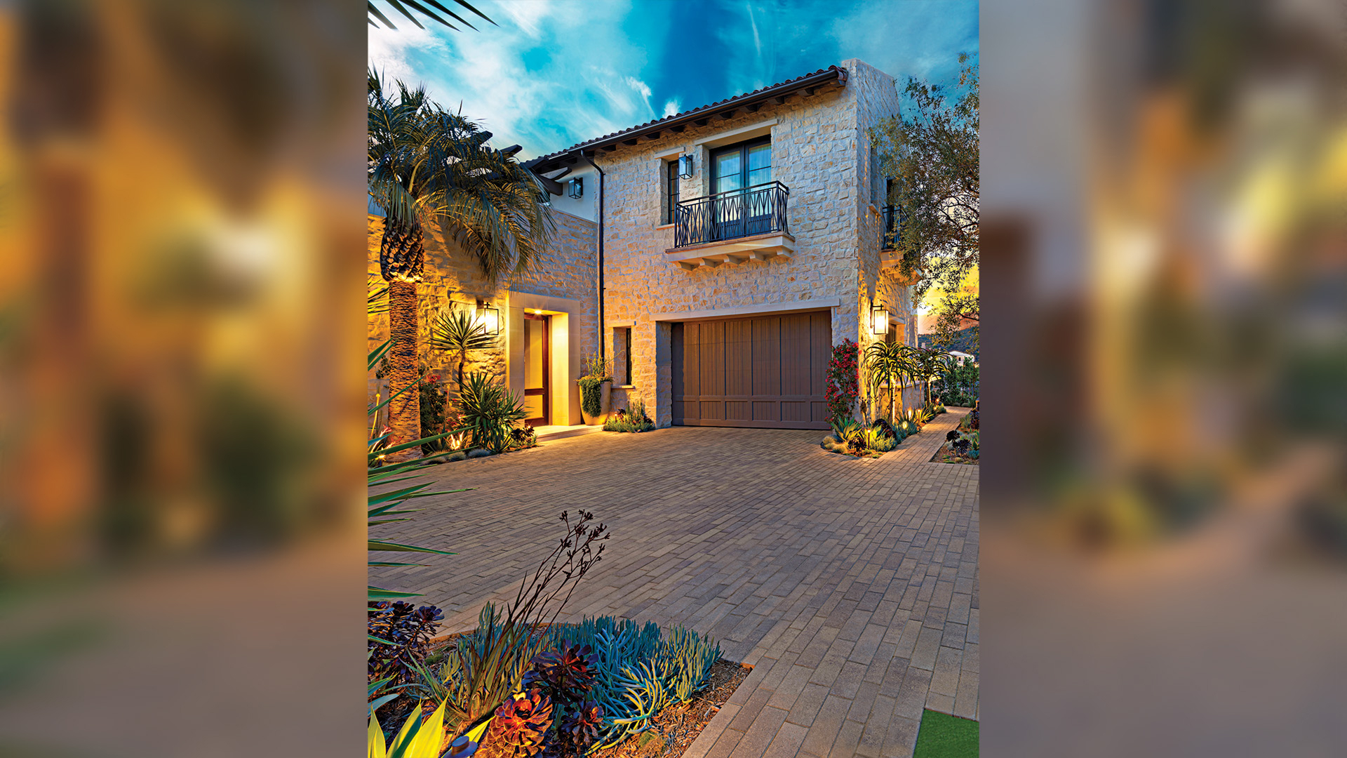 Aqualina pavers in Sand-Stone-Mocha (special order color) installed in a random runner pattern enhance this elegant residential driveway. The smooth linear layout leads up to a stone-faced home with warm lighting and lush tropical landscaping.