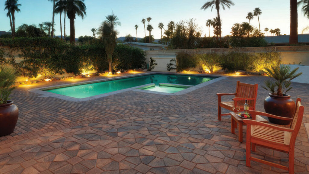 Poolside patio at sunset featuring Cream-Brown-Charcoal Bishops Hat pavers combined with solid Brown 8x8s in a parquet pattern. Near the pool edge, Cream-Brown-Charcoal Holland pavers are installed in a running bond pattern. The space includes wooden chairs, large potted plants, and soft landscape lighting, with palm trees silhouetted against the evening sky.
