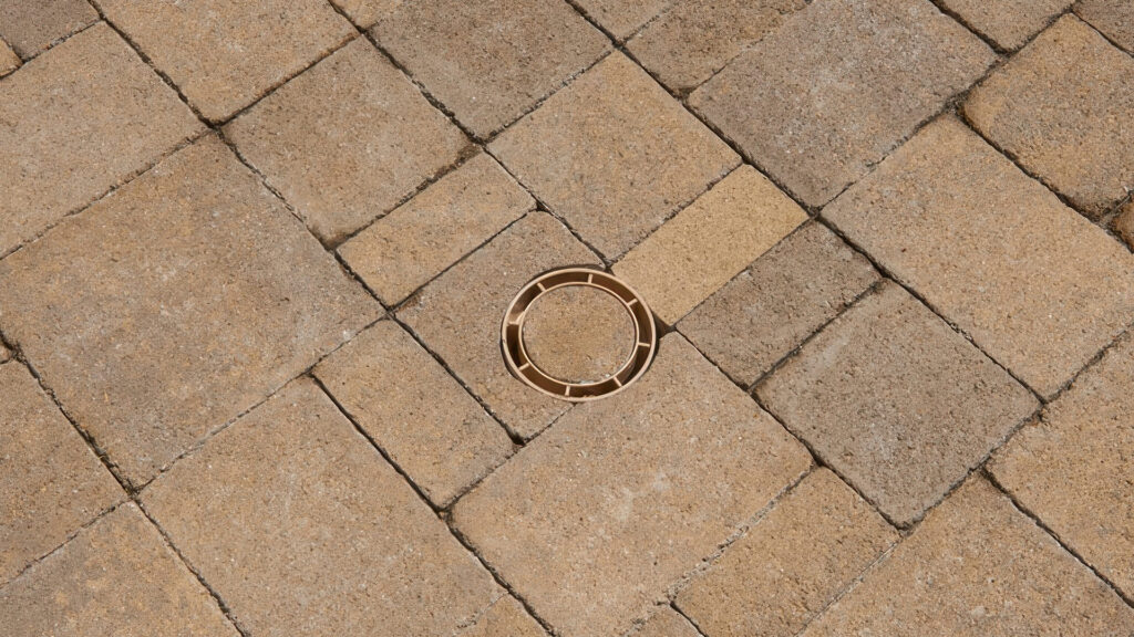 Close-up of a drain integrated into Sand-Stone-Mocha Castle Cobble I and II (distressed) pavers arranged in an ashlar pattern. The drain cover is cleverly camouflaged using the same paving stone material, minimizing its visual impact and blending seamlessly into the surface.