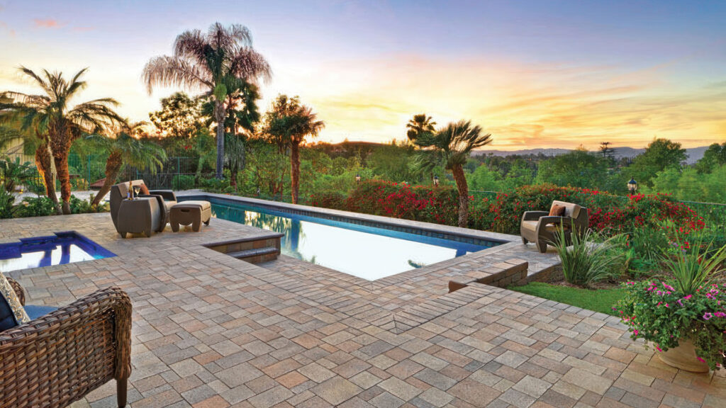 Backyard poolside patio at sunset featuring a 50/50 blend of Cream-Brown-Charcoal and Gray-Moss-Charcoal Castle Cobble I and II (distressed) pavers installed in a random ashlar pattern. The steps and pool coping are finished with solid Charcoal Bullnose, creating a clean transition around the spa and pool. Surrounded by tropical landscaping and contemporary outdoor furniture.