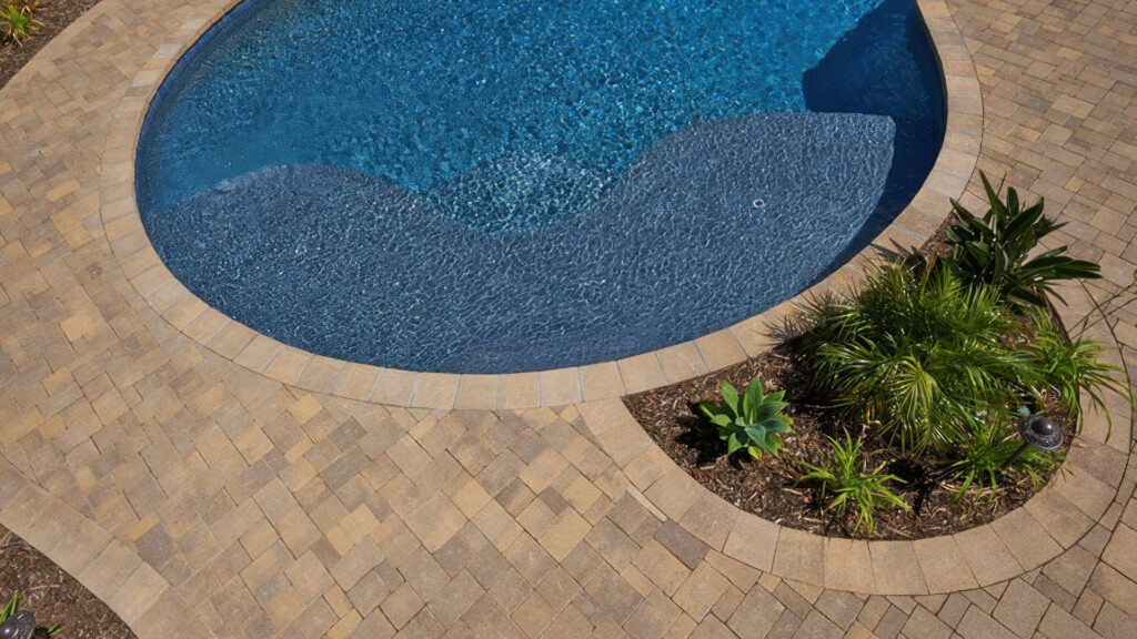 Pool Coping: Distressed Sand-Stone-Mocha Castle Cobble I and II pavers installed in an ashlar pattern surrounding a pool. The pool coping and paver border are made of Sand-Stone-Mocha Castle Cobble II (distressed), forming a clean, curved edge around the pool and adjacent planting area.