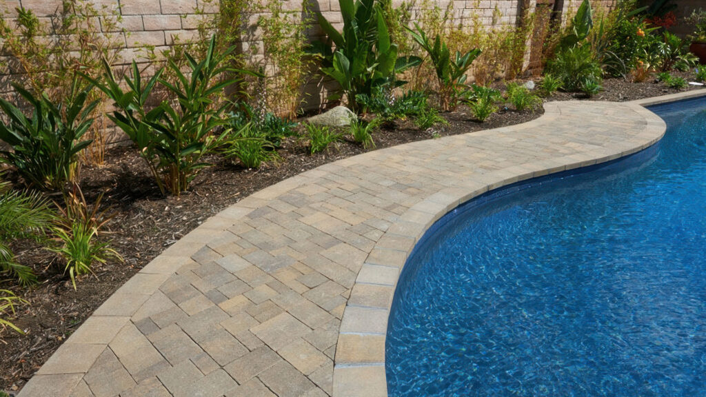 Pool Coping: Distressed Sand-Stone-Mocha Castle Cobble I and II pavers installed in an ashlar pattern surrounding a curved swimming pool. The pool coping and paver border are made from Sand-Stone-Mocha Castle Cobble II (distressed), creating a smooth, elegant transition between the pool and adjacent landscaping.