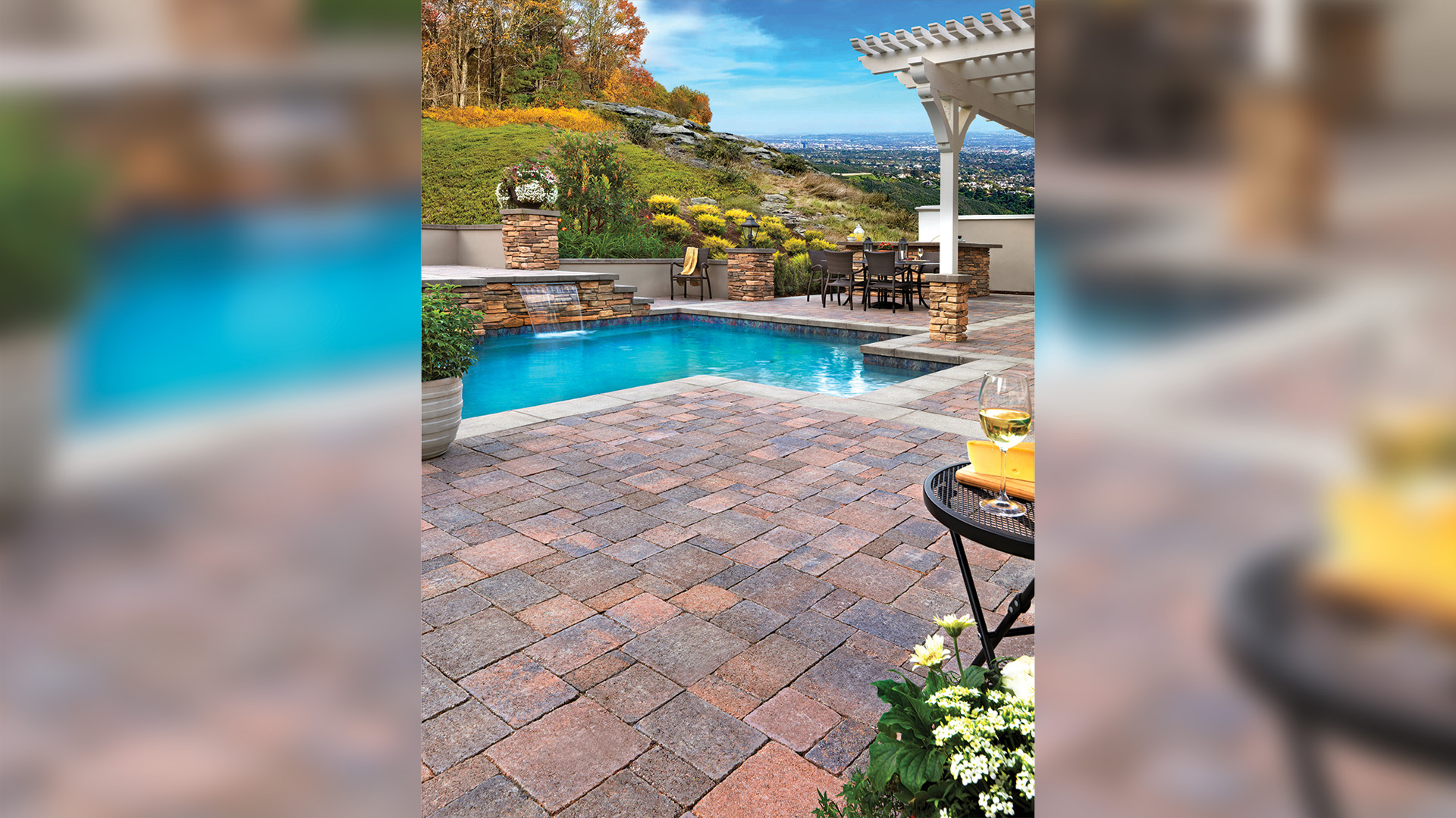 Backyard patio and pool area featuring Cream-Brown-Charcoal Castle Cobble I and II pavers with a distressed finish, installed in a random ashlar pattern. The space includes a rectangular pool with a cascading water feature, a pergola-covered dining area, stone column accents, and colorful landscaping with a scenic hillside view in the background.