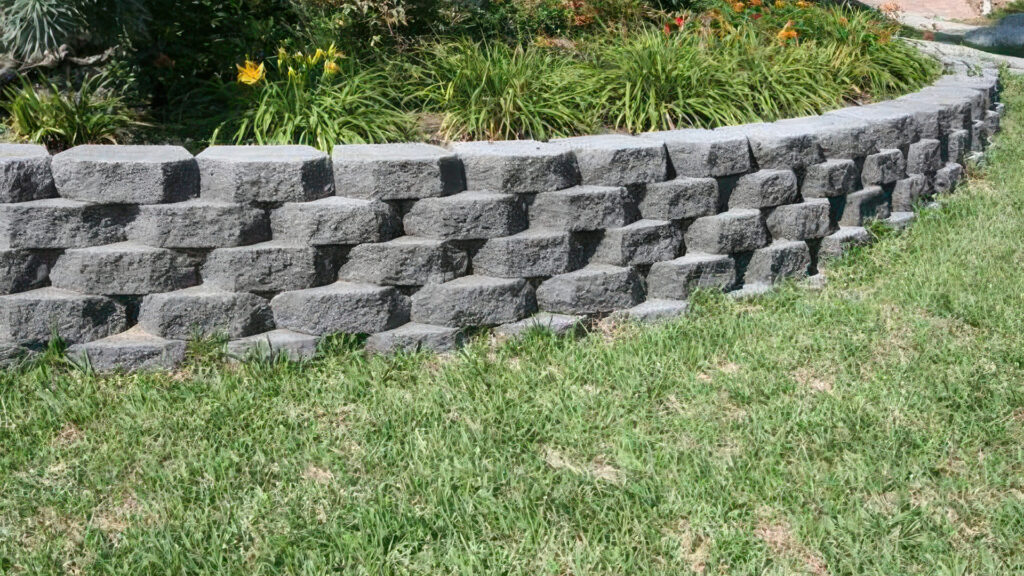 12-inch Charcoal planter wall constructed in a curved formation with staggered, textured blocks. The wall borders a raised garden bed filled with green ornamental grasses and blooming yellow and orange daylilies, set against a well-maintained lawn.