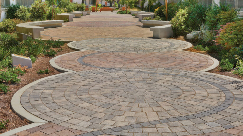Decorative pedestrian walkway featuring interlocking Courtyard Circles in multiple color blends, including shades of gray, tan, and red. The circular patterns are bordered by curved concrete edging and surrounded by lush landscaping. Concrete benches line the pathway, leading to a central gathering area with additional seating and ornamental trees.