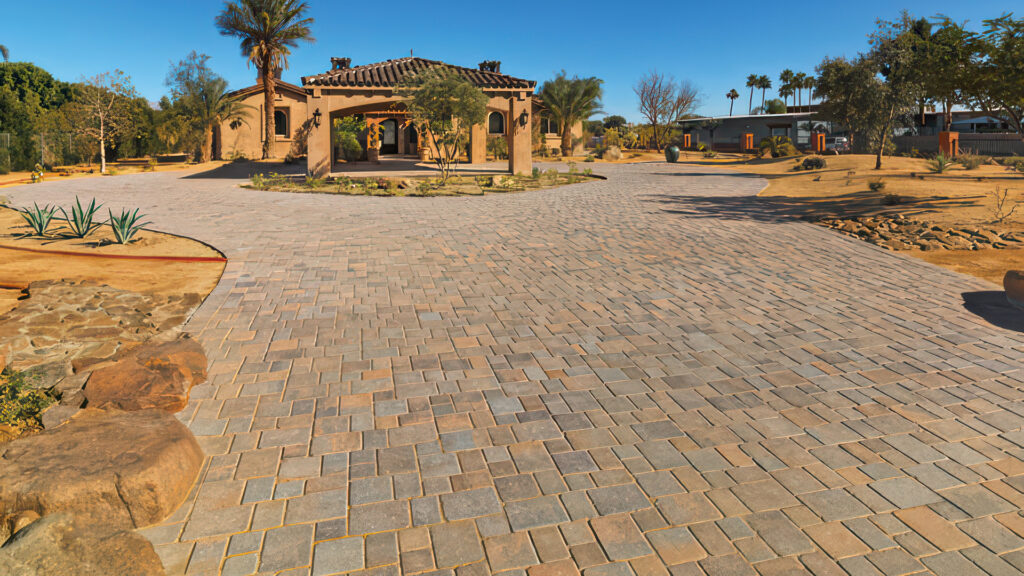 Expansive driveway paved with Tuscan Courtyard Stone (tumbled) in an '8 pattern', leading to a Mediterranean-style home surrounded by a desert landscape with rocks, native plants, and palm trees under a clear blue sky.