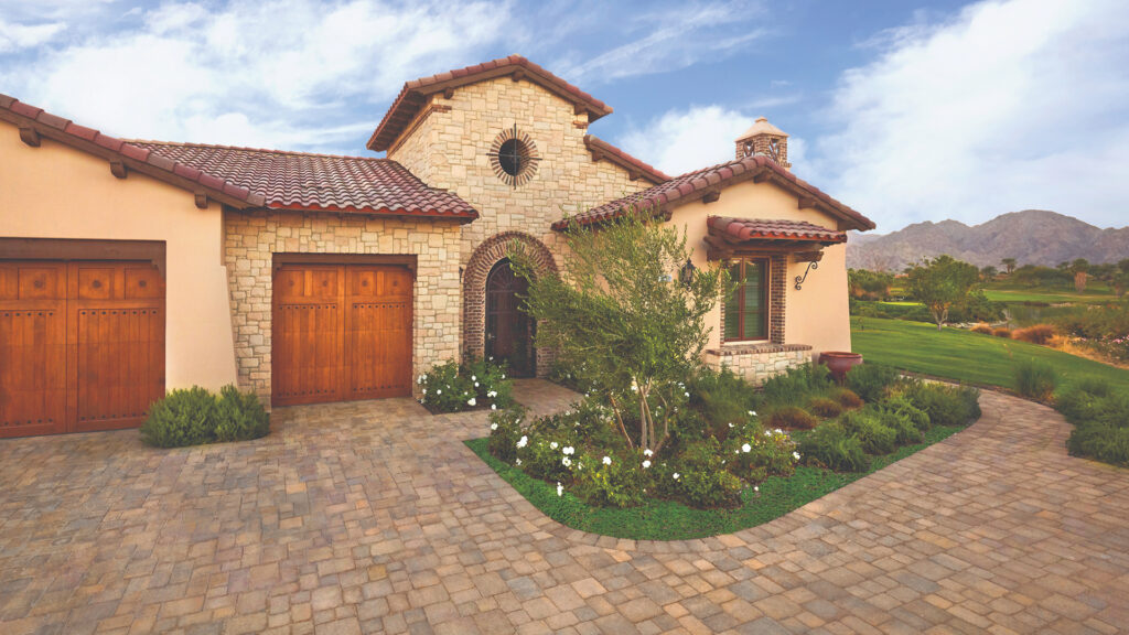 Front view of a Mediterranean-style home with a tumbled Sand-Stone-Mocha Courtyard Stone driveway and walkway, installed in a random ashlar pattern. The warm-toned paving stones complement the stone façade, wooden garage doors, and landscaped garden with white flowers and desert plants. A scenic mountain range is visible in the background.