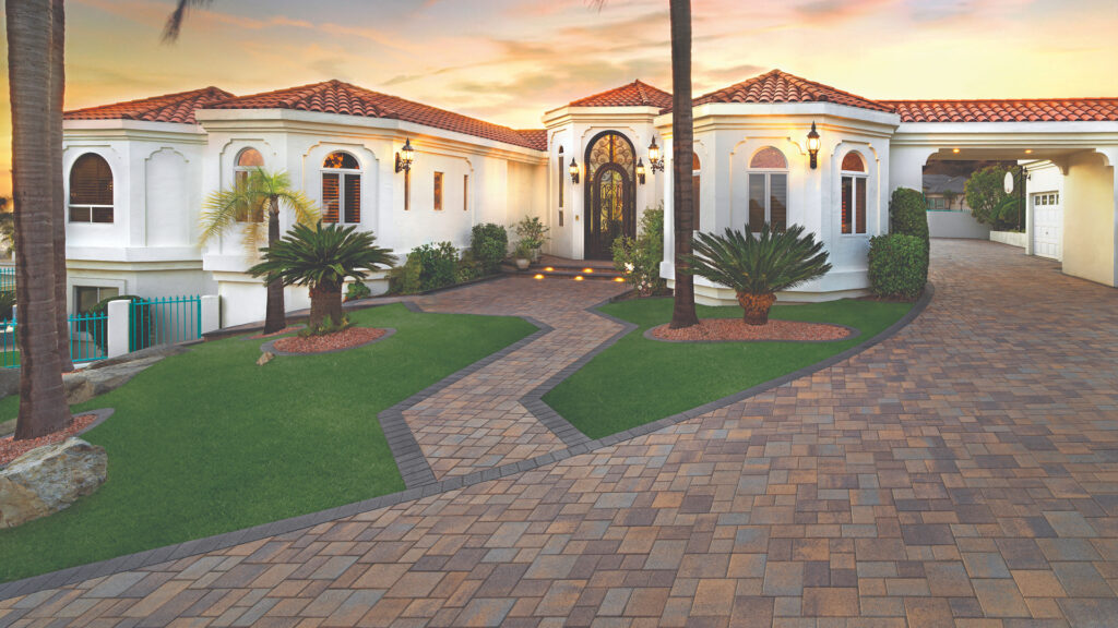 Driveway and walkway paved with Courtyard Stone in Tuscan Blend, installed in a random ashlar pattern. Pathway is bordered with solid Charcoal Courtyard 6x9 stones in a soldier course. Mediterranean-style home features white stucco, red tile roof, and manicured landscaping with palm trees and green turf.