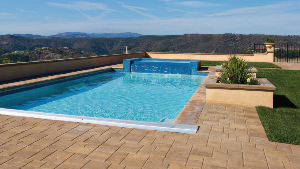 Backyard pool area with a scenic mountain view, featuring Sand-Stone-Mocha Courtyard Stone pavers installed in an ashlar pattern. The space includes a rectangular pool with a raised spa wall covered in blue tile, manicured lawn, built-in planters, and cream-colored walls that frame the patio.