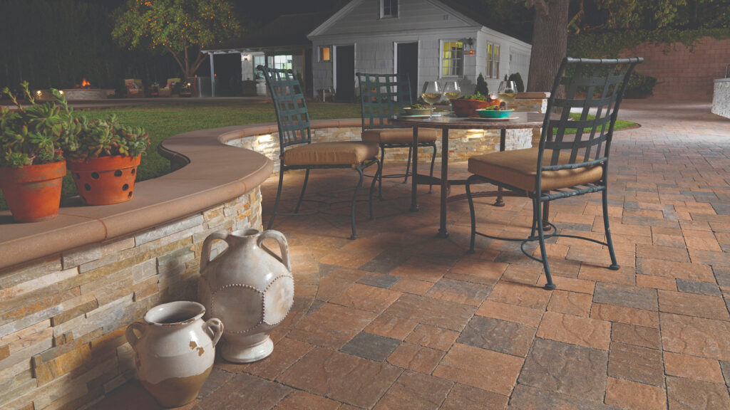 Outdoor dining space with a curved stone seating wall and rustic patio set, featuring Cream-Brown-Charcoal Slate Stone pavers arranged in an ashlar pattern, creating a warm and textured surface ideal for evening gatherings.
