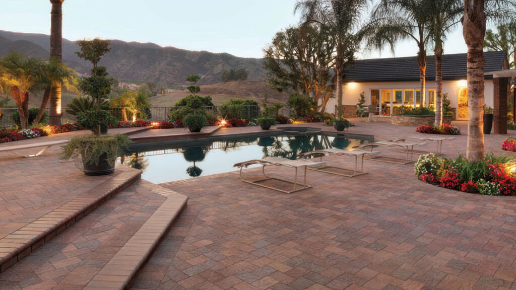 Expansive backyard patio with a pool surrounded by 6x9 Cream-Brown-Charcoal Slate Stone pavers installed in a herringbone pattern. The space features built-in steps with matching pavers, multiple lounge chairs, vibrant flower beds, palm trees, and soft landscape lighting set against a scenic hillside backdrop at dusk.