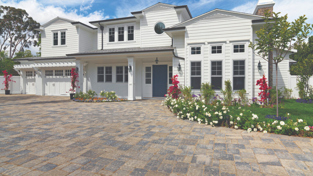 Driveway paved with Estate Cobble I and II in Gray–Moss–Charcoal blend, installed in a tumbled ashlar pattern. The bright white two-story home features black-trimmed windows, a navy blue front door, and vibrant landscaping with colorful flowers and lush greenery.