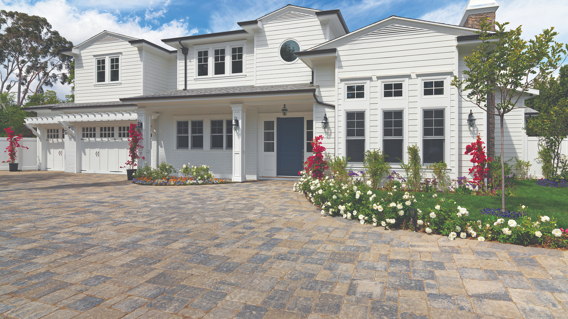 Driveway paved with Estate Cobble I and II in Gray–Moss–Charcoal blend, installed in a tumbled ashlar pattern. The bright white two-story home features black-trimmed windows, a navy blue front door, and vibrant landscaping with colorful flowers and lush greenery.