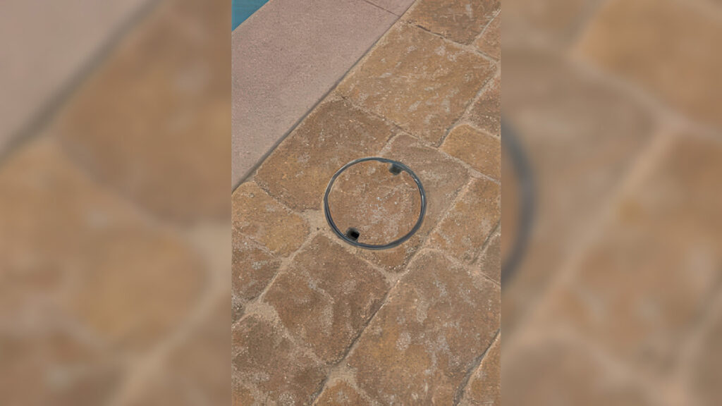 Close-up of a drain cover set into tumbled Sand-Stone-Mocha Estate Cobble I and II pavers, arranged in an ashlar pattern. The paving surrounds a pool deck area, showcasing the rustic texture and warm earth tones of the cobblestones.