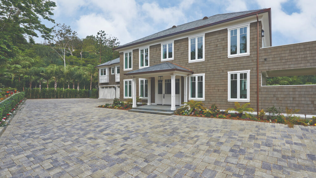 Spacious driveway paved with Estate Cobble I and II in Gray–Moss–Charcoal blend, installed in a tumbled ashlar pattern. The two-story home features taupe shingle siding, white trim, and a landscaped border with colorful flowers and lush greenery.