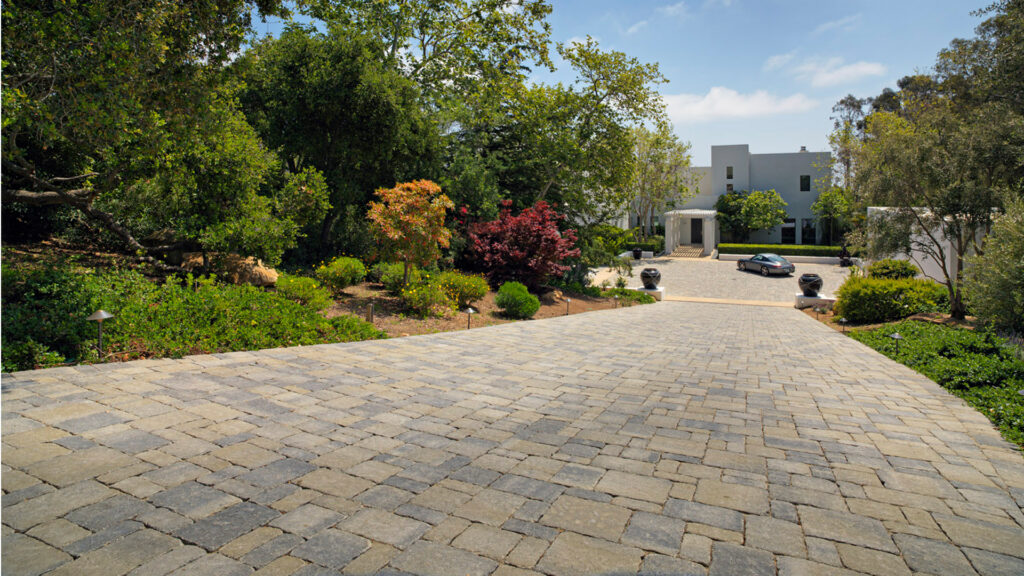 Long driveway paved with tumbled Estate Cobble I & II in Gray–Moss–Charcoal blend, installed in a random ashlar pattern. The setting features lush, mature landscaping and leads to a modern white estate with circular motor court.