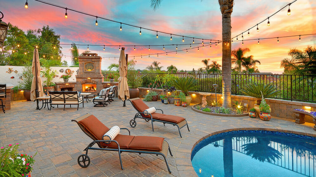 Vibrant backyard patio featuring Estate Cobble I and II pavers in Tuscan color, installed in a random ashlar pattern. The space includes a cozy fireplace seating area, lounge chairs by a curved-edge pool, colorful potted plants, and hanging string lights overhead. Tropical landscaping and a warm sunset sky complete the inviting atmosphere.