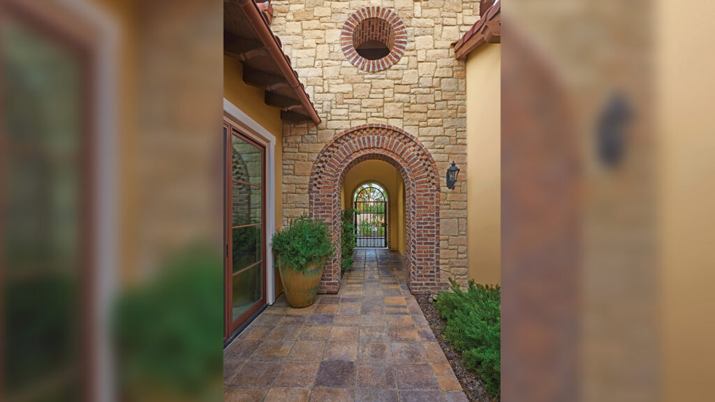 Mediterranean-style entryway featuring Sand-Stone-Mocha Estate Cobble II pavers arranged in a stack bond pattern. The walkway leads through a brick-accented stone archway with a wrought iron gate at the far end. Surrounding the walkway are warm stucco walls, clay roof tiles, and potted greenery.