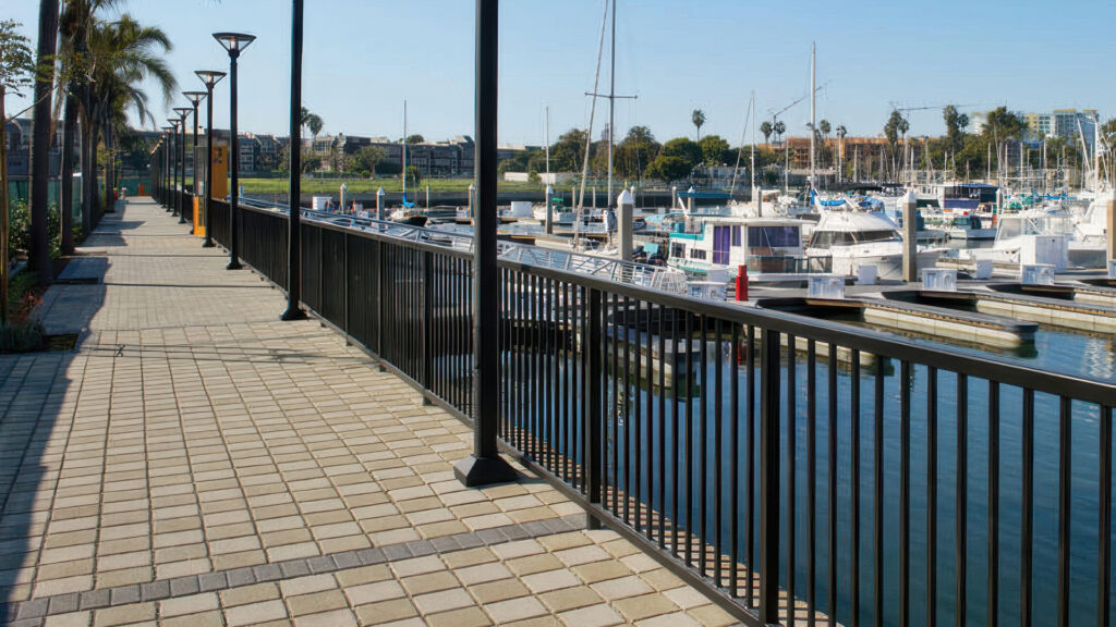 Waterfront pedestrian promenade with a black metal railing overlooking a marina filled with yachts and sailboats. The walkway is paved with SF Rima pavers in a 50/50 blend of Gray and Moss, arranged in a clean stack bond pattern. Modern lamp posts line the path, and palm trees provide shade on the opposite side, enhancing the coastal atmosphere.