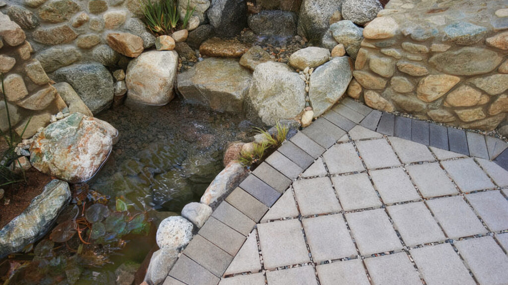 Close-up of a natural stone water feature with a gentle waterfall flowing into a small pond, adjacent to a permeable patio made of Gray SF Rima pavers. The paving includes a Gray-Moss-Charcoal Holland stone border in a running pattern, designed to allow water to soak into the ground for effective drainage.