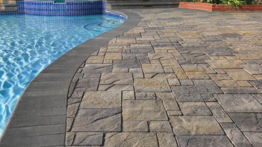 Poolside patio featuring Gray-Moss-Charcoal Heartland Pavers installed in an ashlar pattern. The curved pool edge and steps are finished with Charcoal bullnose coping, complementing the textured surface of the pavers. The scene includes clear blue pool water and a raised spa with blue tile facing.