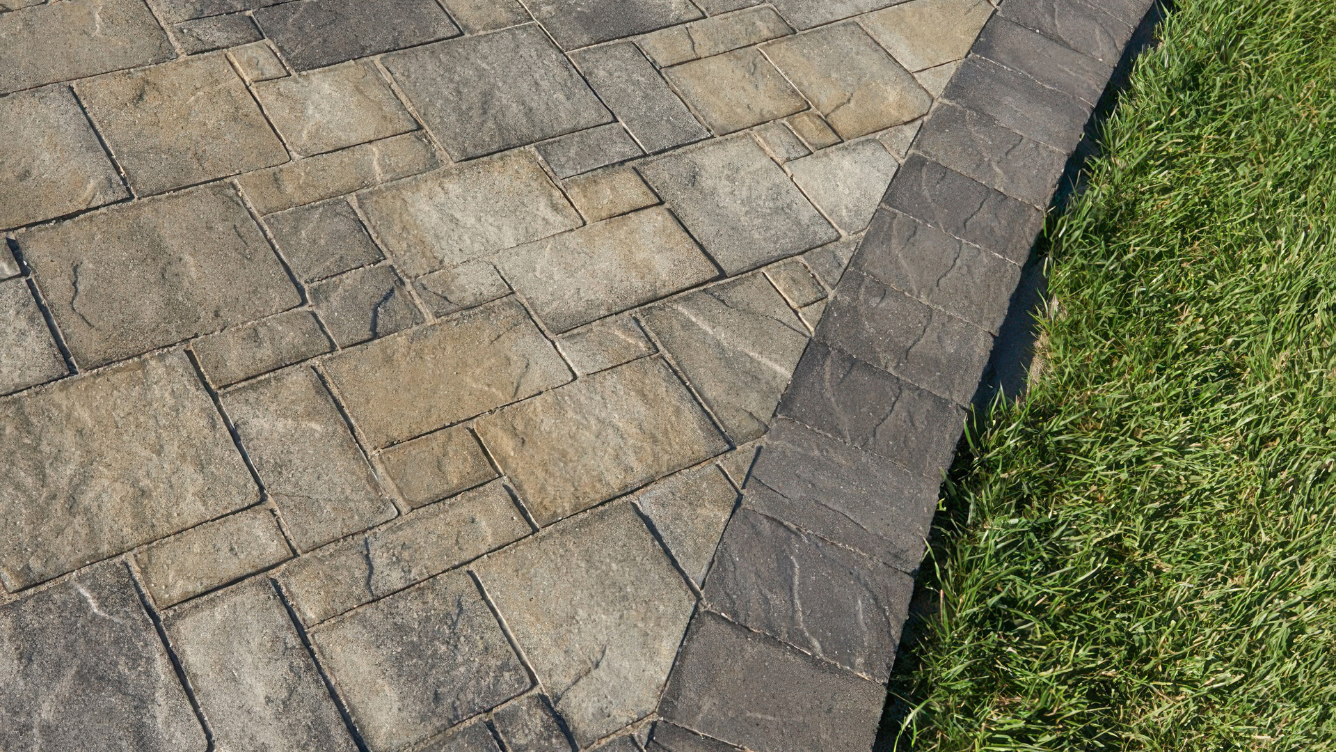 Gray-Moss-Charcoal Heartland Stone pavers installed in an ashlar pattern. The border features a clean soldier course of solid Charcoal Slate Stone 6x9 (distressed), curving gently alongside a lush green lawn.