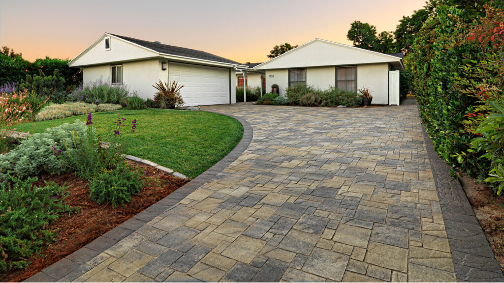 Curved residential driveway paved with Heartland paving stones in Gray–Moss–Charcoal blend, bordered with darker accent pavers. The setting features a single-story white home, lush green lawn, and vibrant landscaping with flowering plants and shrubs.