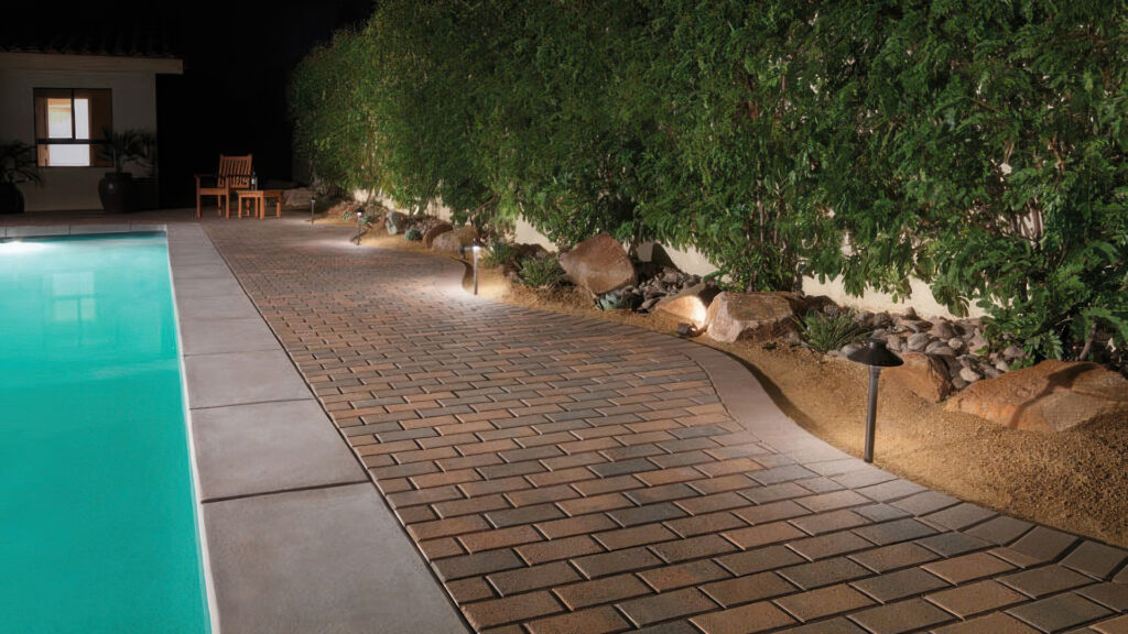 Evening view of a poolside walkway featuring Cream-Brown-Charcoal Holland pavers installed in a running bond pattern. The path is bordered with a solid Brown Holland paver edge along the landscaped area. Landscape lighting softly illuminates the walkway, which is flanked by greenery, decorative boulders, and a seating area near the house.