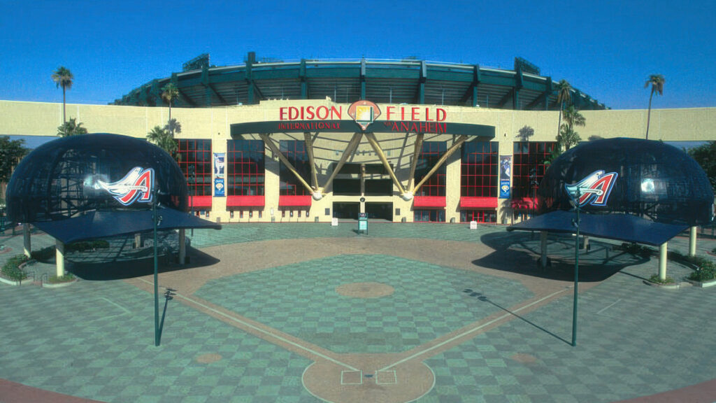 Entrance plaza of Edison Field in Anaheim, featuring two oversized batting helmet sculptures flanking the main gate. The ground is paved with Holland pavers in a checkerboard layout using Cream and Gray, along with custom colors including Dark Red, Dark Green, Light Green, Gold, Light Gold, and Burgundy. The paver design includes a stylized baseball diamond embedded into the surface, adding thematic detail to the stadium entrance.