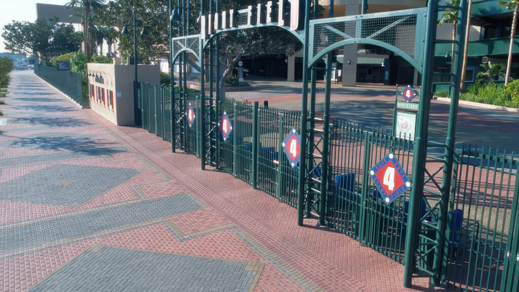 View of a stadium entrance marked as Gate 4 with green metal fencing and an arched sign. The walkway is paved with Holland pavers in a mix of Cream and Gray, enhanced by custom colors including Dark Red, Dark Green, Light Green, Gold, Light Gold, and Burgundy. The pavers are arranged in detailed geometric patterns, including diamonds and borders, adding visual interest to the stadium’s exterior plaza.