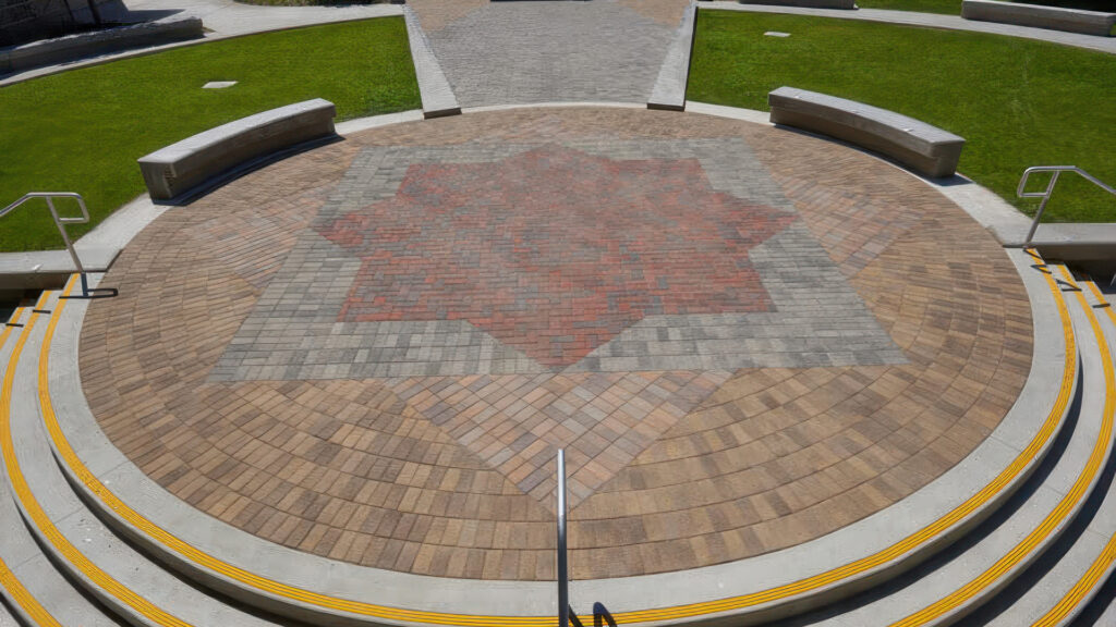 Circular amphitheater paving features an intricate pattern of Holland stones, showcasing color blends of Sand-Stone-Mocha, Tuscan, Gray-Moss-Charcoal, and Red-Brown-Charcoal. The bold star design in the center is made with Red-Brown-Charcoal, bordered by Gray-Moss-Charcoal, and framed with surrounding circular bands in Sand-Stone-Mocha and Tuscan. Concrete stairs with yellow safety treads lead down to the paved area, flanked by curved benches and green lawns.