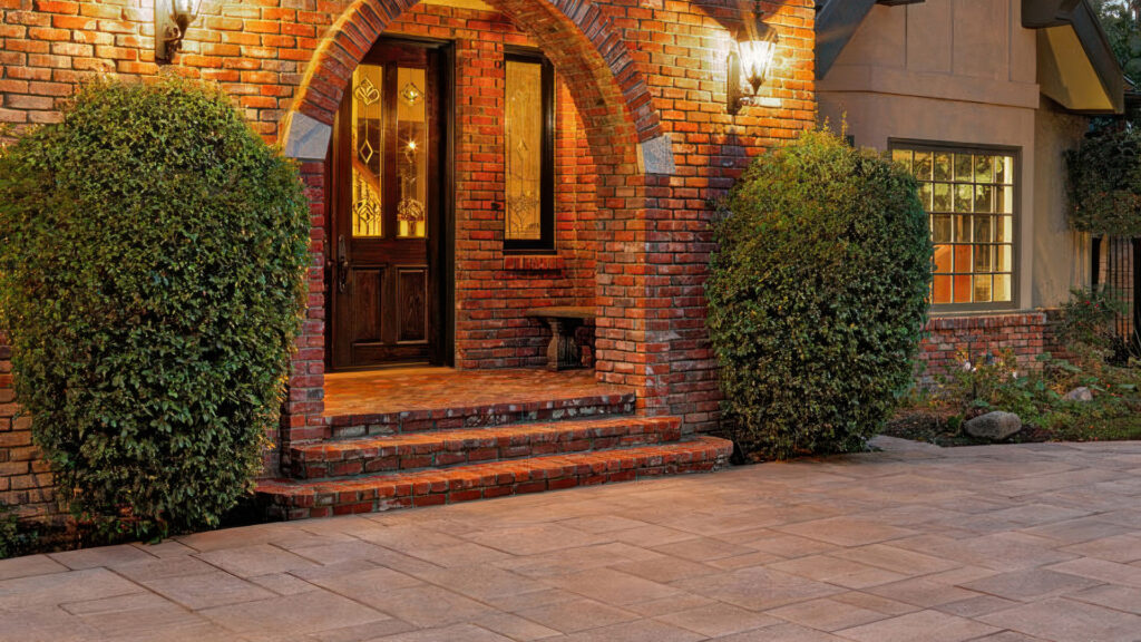 Paseo I and II paving stones in an Adobe-Copper-Mocha blend laid in an ashlar pattern, creating a warm-toned front entryway. The paving complements the red brick façade, arched doorway, and lantern-style wall sconces, framed by manicured shrubs on each side.