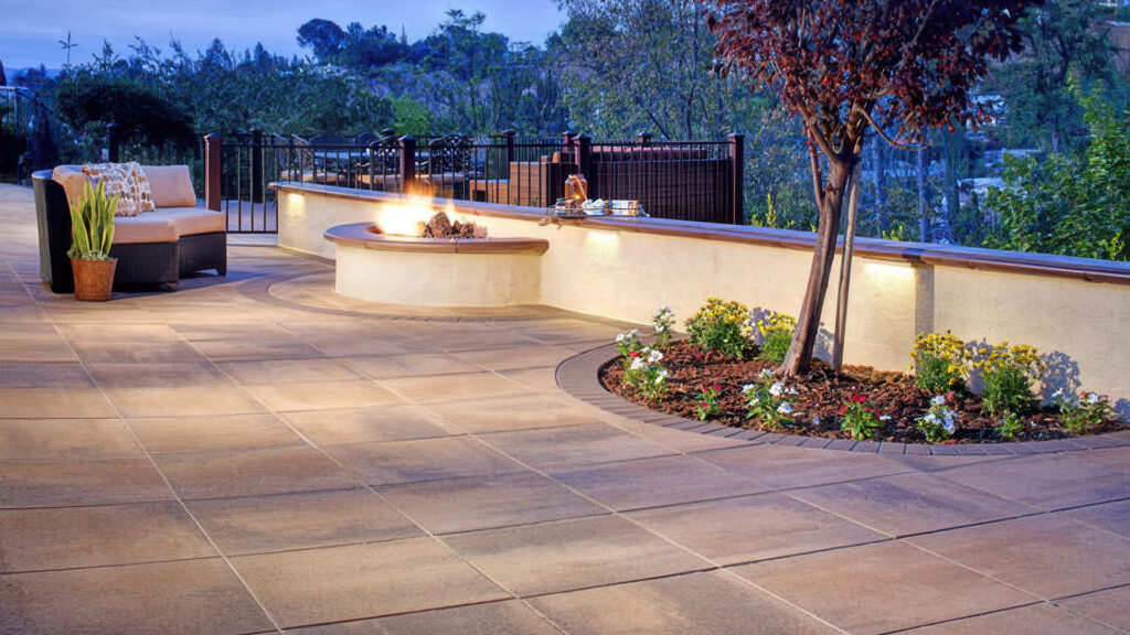 Outdoor patio paved with Paseo II pavers in Adobe-Copper-Mocha, featuring a smooth, elegant layout. The curved flower bed border is defined with solid Mocha Holland stone in a soldier course. A built-in stucco bench with integrated fire feature and ambient lighting adds warmth and comfort to the landscaped space.