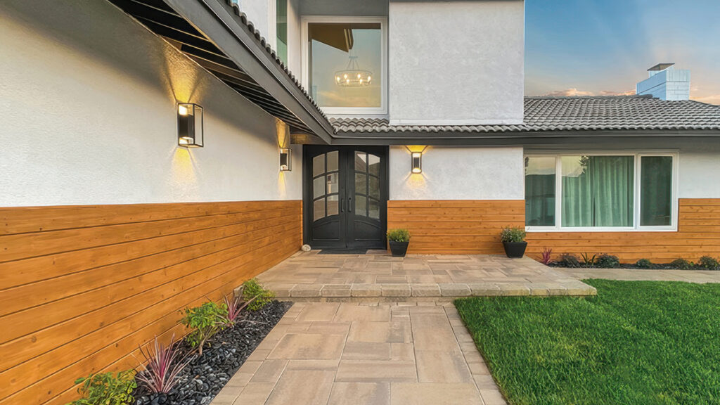 Front entryway featuring Adobe-Copper-Mocha Paseo I & II pavers in an ashlar pattern, leading to a dark double door. The raised step is made from tumbled Sand-Stone-Mocha Estate Cobble II. The surrounding design includes warm wood siding, black light fixtures, and a lush green lawn.